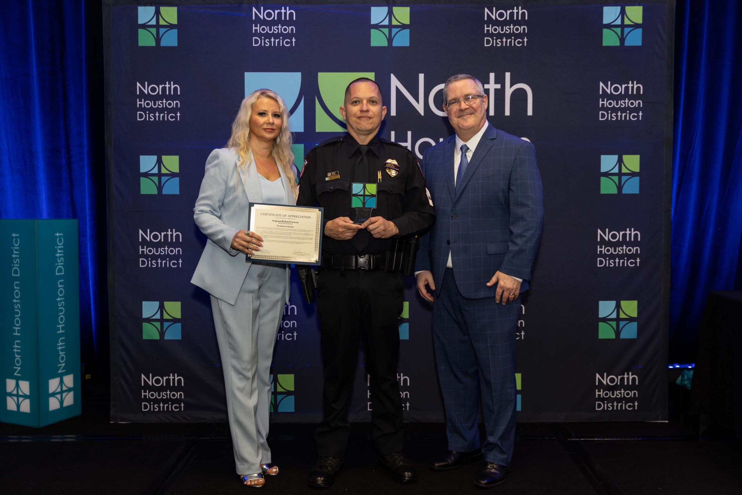 Three people stand together smiling. A woman in a blue suit holds a certificate with a police officer in uniform, while a man in a blue plaid suit stands to the side.