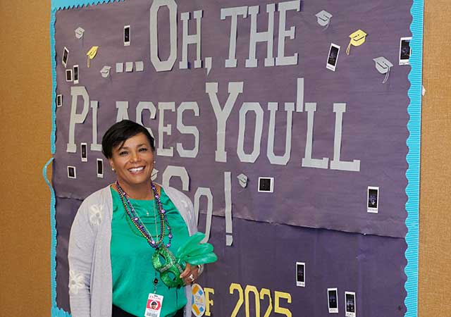 A woman stands smiling in front of a bulletin board that says Oh, the Places You’ll Go! with photos of graduates.
