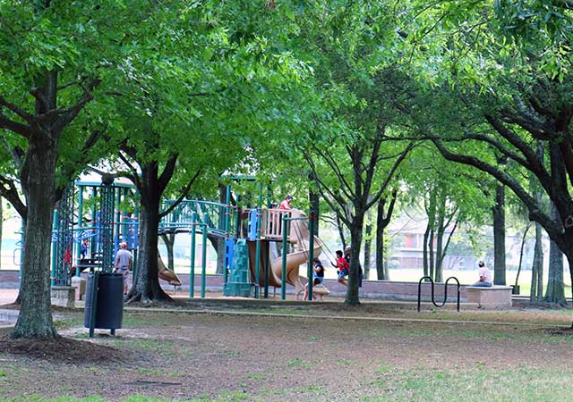 A wide view of Wussow Park shows a shaded playground surrounded by mature trees, with children playing on equipment and open green space extending through the park.