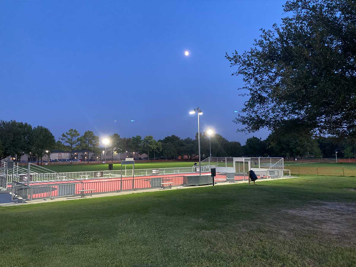 Evening view of the brightly lit mini soccer field at Thomas R. Wussow Park, with surrounding trees, open grassy space, and the moon visible in the clear night sky.