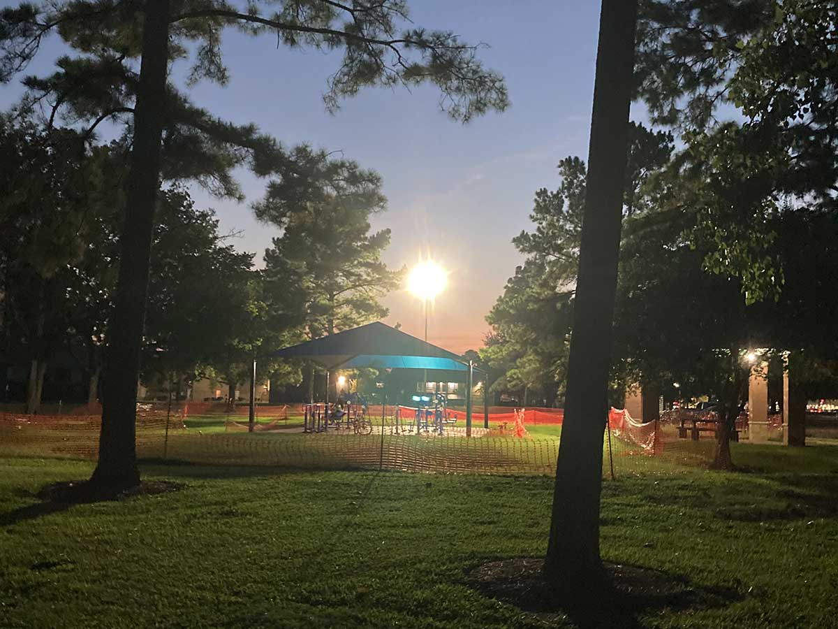 Playground area at Thomas R. Wussow Park illuminated at dusk, with a blue shade canopy, construction fencing, and tall trees creating a peaceful park setting.