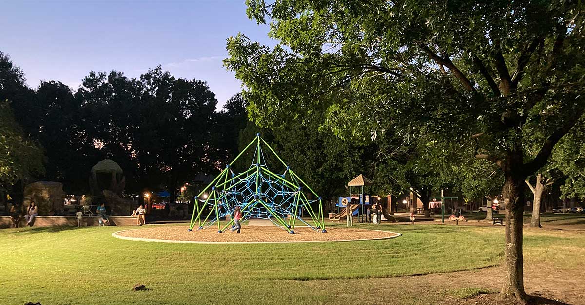 Wussow Park Renovation Newsroom Graphic Evening view of the playground at Thomas R. Wussow Park with children playing on a large illuminated climbing structure under tall trees and a darkening sky.