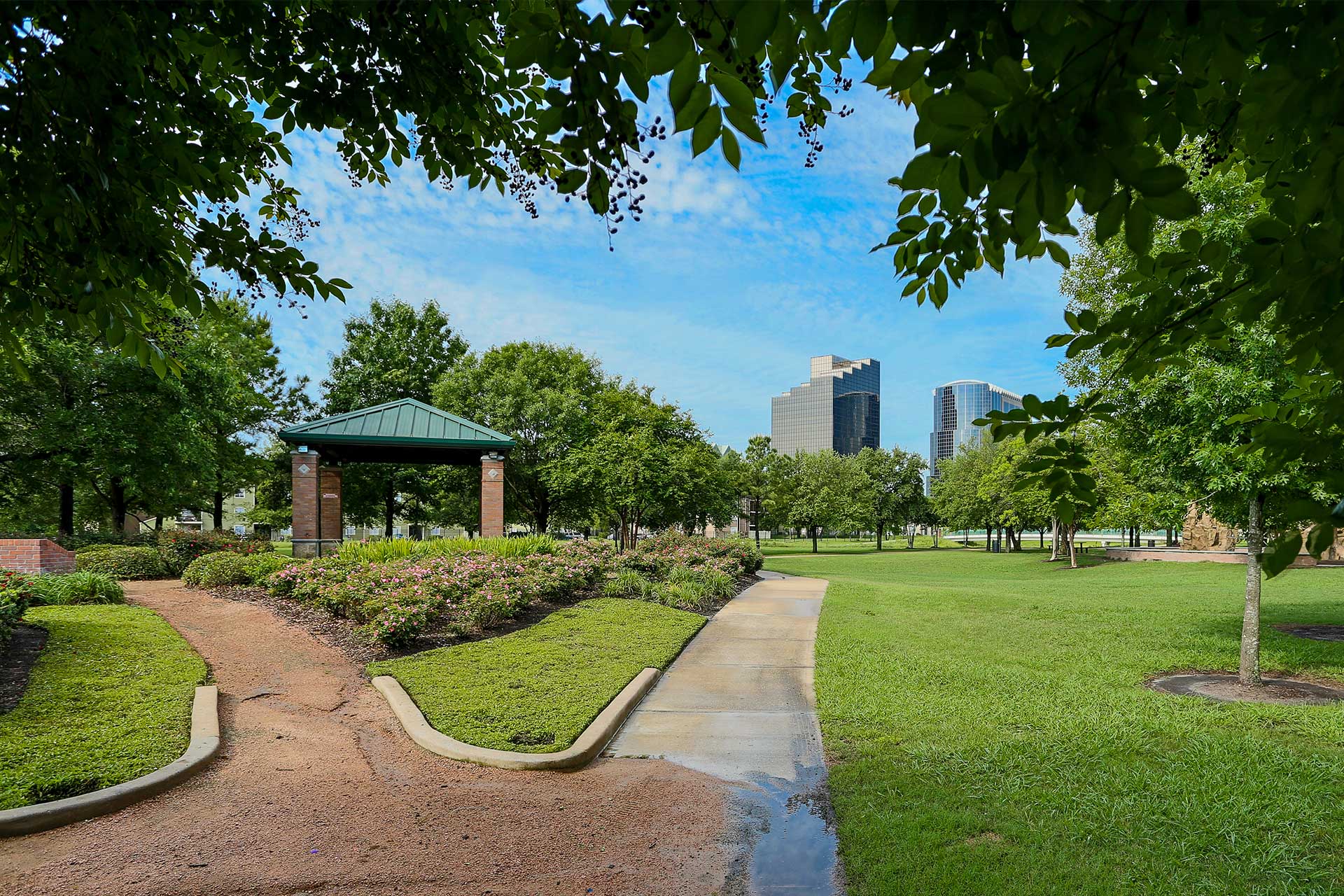 This image shows a peaceful park scene at Wussow Park with a gazebo in the center, framed by green trees and well-manicured shrubs. A paved walkway leads to the gazebo, and a parallel gravel path curves around it. In the background, several tall, modern skyscrapers rise above the treetops, contrasting with the natural setting of the park. The sky is bright blue with light clouds, and the area appears lush and green.