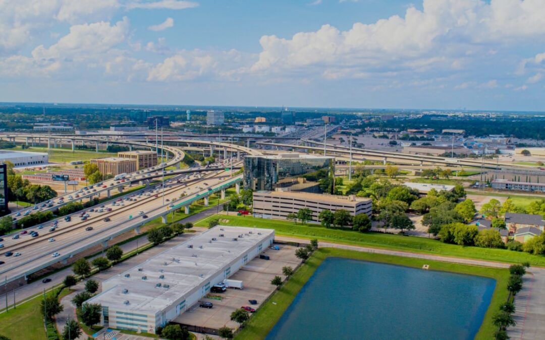 Aerial drone view of a large logistics and distribution center located near a major freeway. The image shows several expansive warehouses with trucks parked at loading docks. The freeway, busy with traffic, runs alongside the facility, with clear blue skies and bright sunlight highlighting the buildings and surrounding roads. Green spaces and other industrial facilities are visible in the background, indicating a well-developed commercial